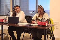 [ai] Two women sitting at a table with a laptop and papers during a discussion in a well-lit room. Large windows provide a view of a city skyline in the background.